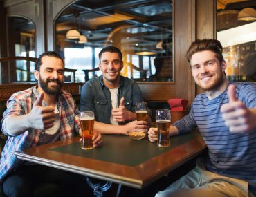 Happy male friends drinking beer and showing thumbs up at bar